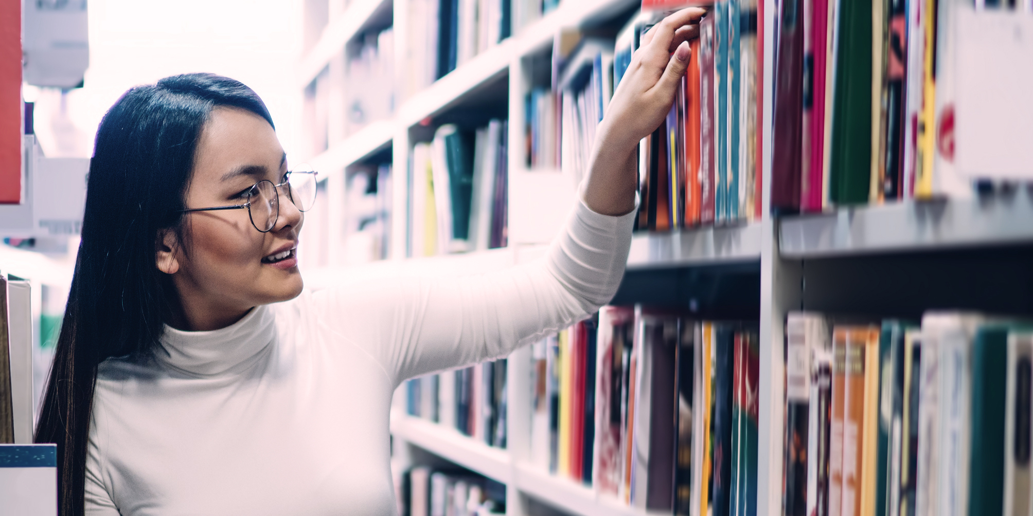 A woman with black hair and glasses selects a book from a library shelf.