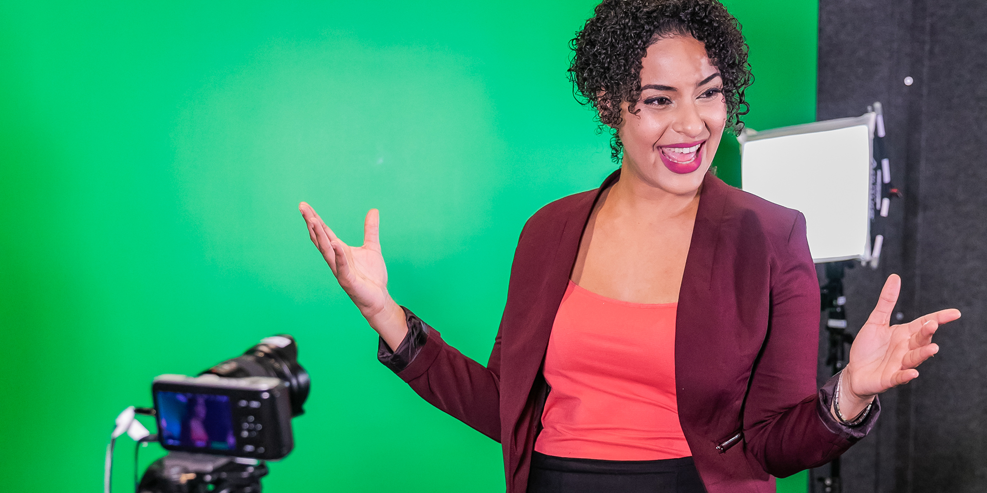 A woman wearing a suit stands inside a recording studio. She poses with her hands up in front of a green screen, microphone and video camera.