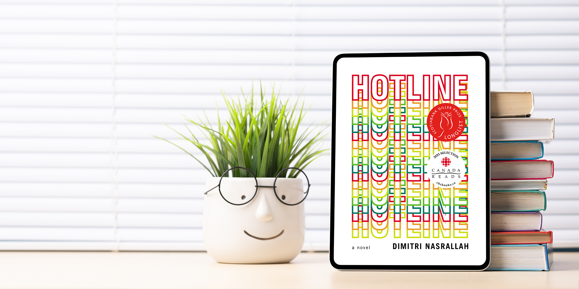 Standing on a wooden table, a tablet shows the book cover for Hotline by Dimitri Nasrallah, which features the word Hotline cascading down the cover. A stack of books props the tablet up. A small plant with a ceramic face wearing glasses sits next to the tablet.